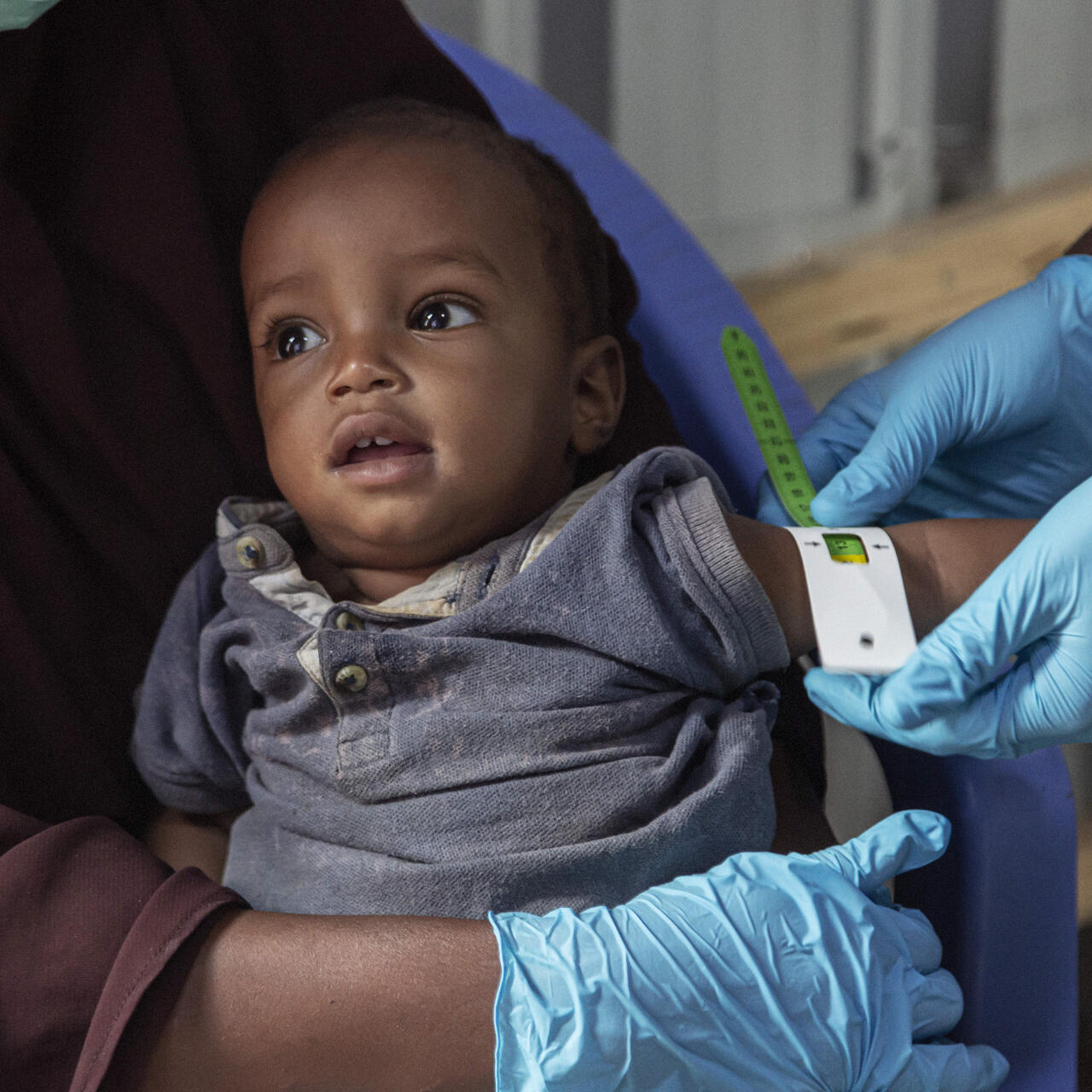 Amina's son, while sitting on his mother's lap, has his arm circumference measured by an IRC staff member wearing blue gloves.