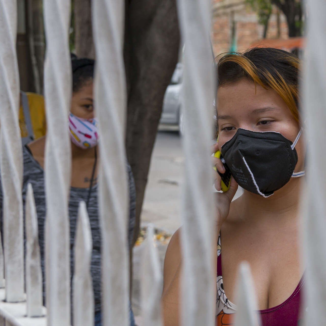 Women wearing face masks wait alongside a fence to enter an IRC health clinic on the Colombia/Venezuela border. One talks on her mobile phone.