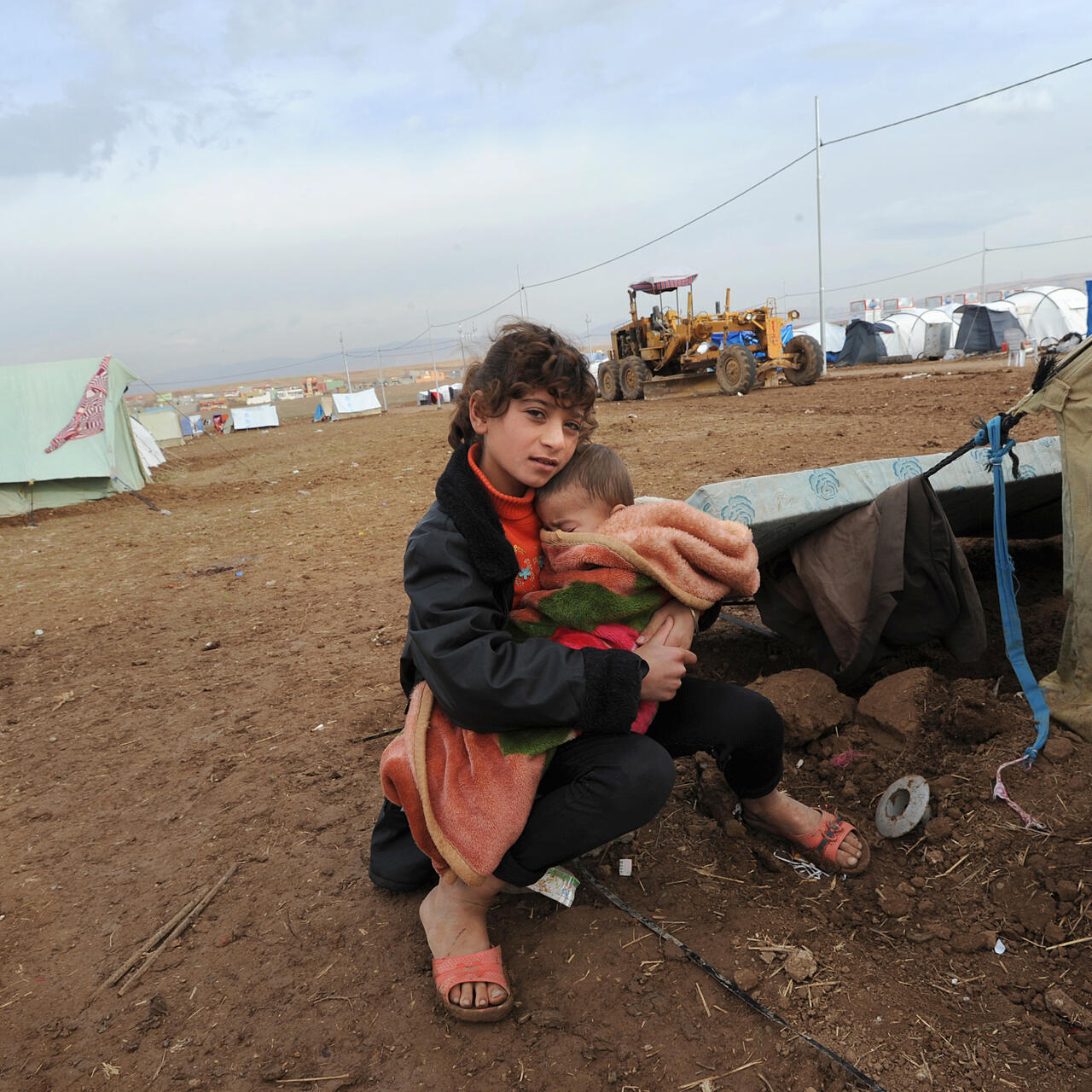 A displaced girl sits just outside a tent in Dohuk, Iraq holding a young child in her arms, bundled in a blanket.