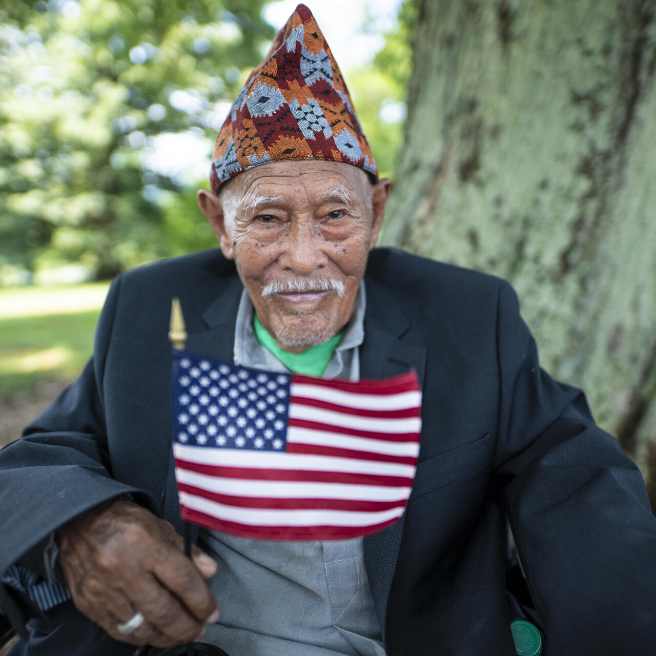 An older man and refugee at a citizenship ceremony sits in front of a tree holding an American flag
