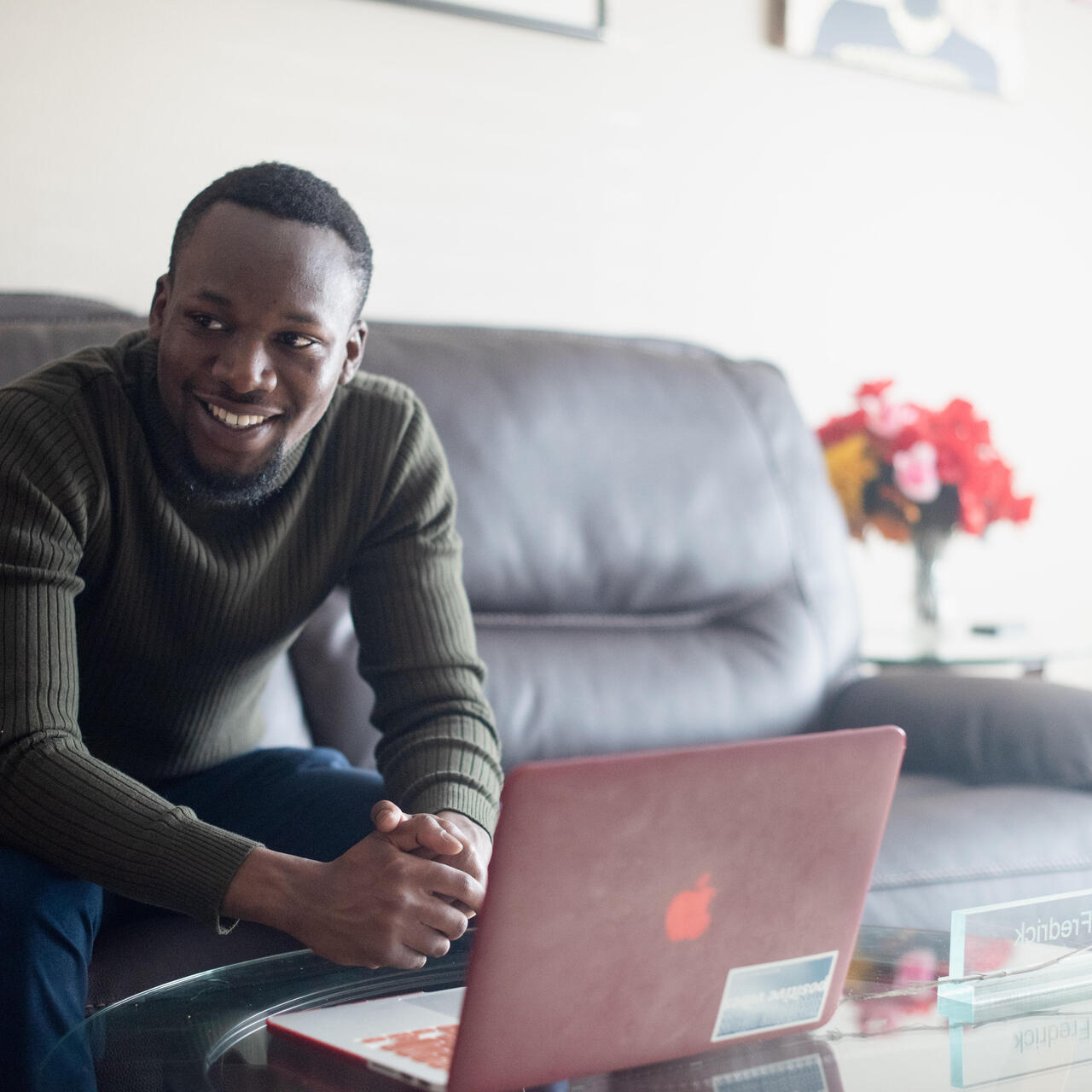Fredrick sits on a couch in his living room with a laptop on a table in front of him. He is looking at the camera and smiling. 