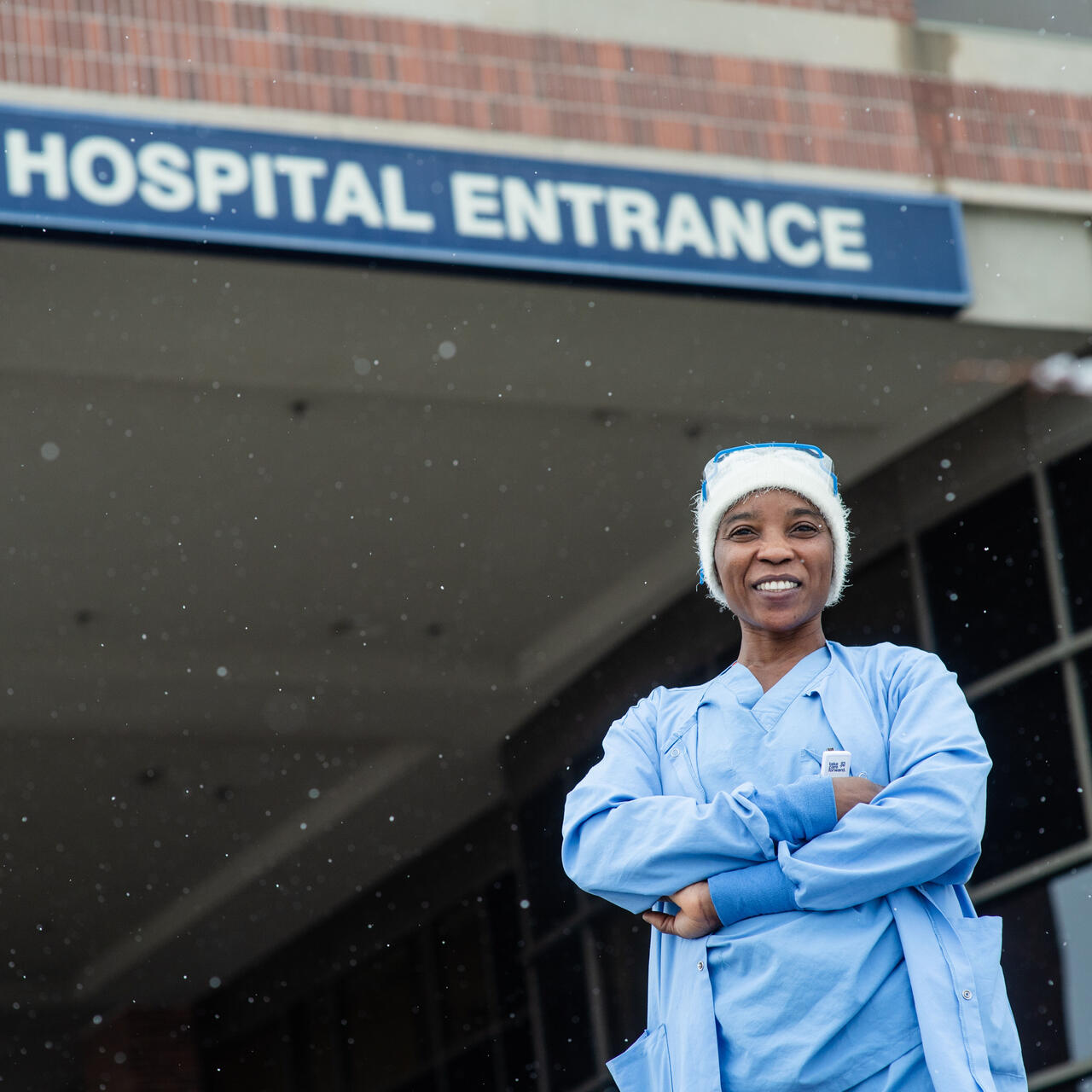 Nabila stands with her arms crossed and smiling in front of the hospital where she works. She is wearing blue scrubs and a white winter hat.