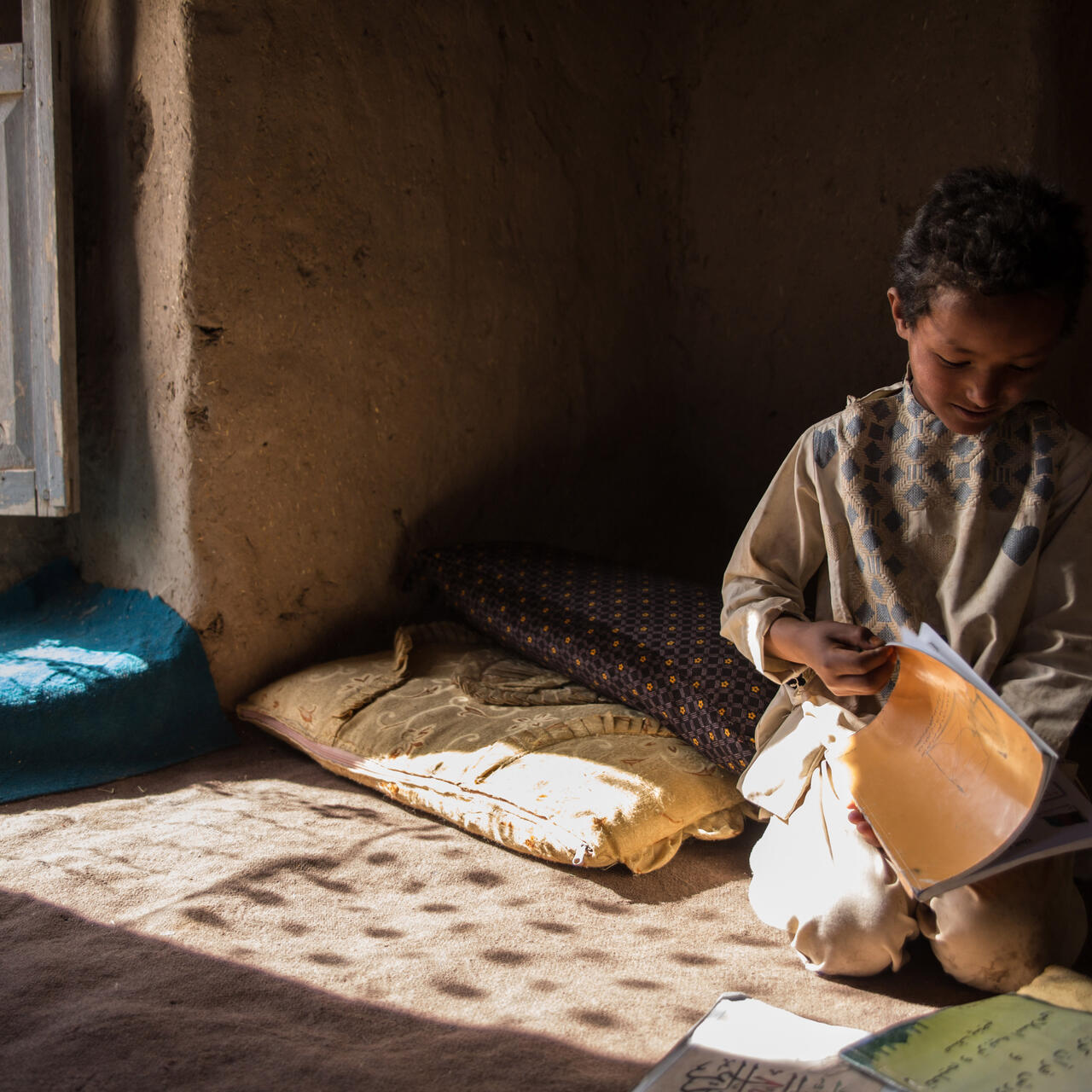 A young boy sits on a mat reading a school exercise book in his family's home in Afghanistan.