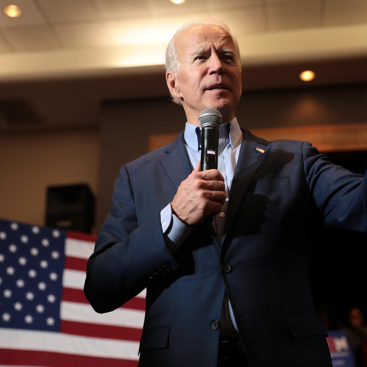 President Joe Biden speaks into a microphone in his hand and gestures with his other hand while standing in front of an American flag