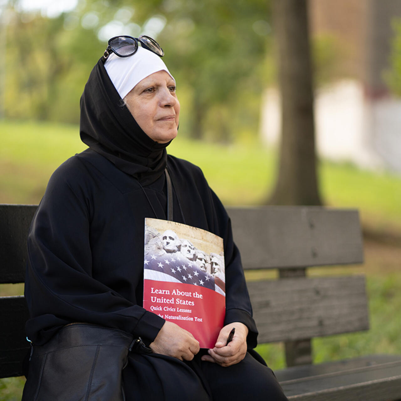 Maha al-Obaidi sits on a park bench looking off into the distance and holding a U.S. citizenship test study book. 