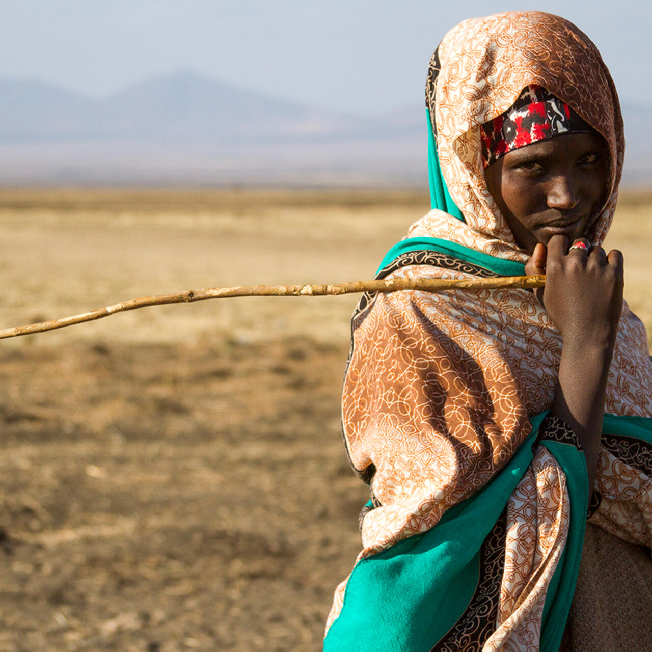 A teenage girl, holding a stick and looking at the camera, stands in a drought affected very dry landscape with mountains in the distance.