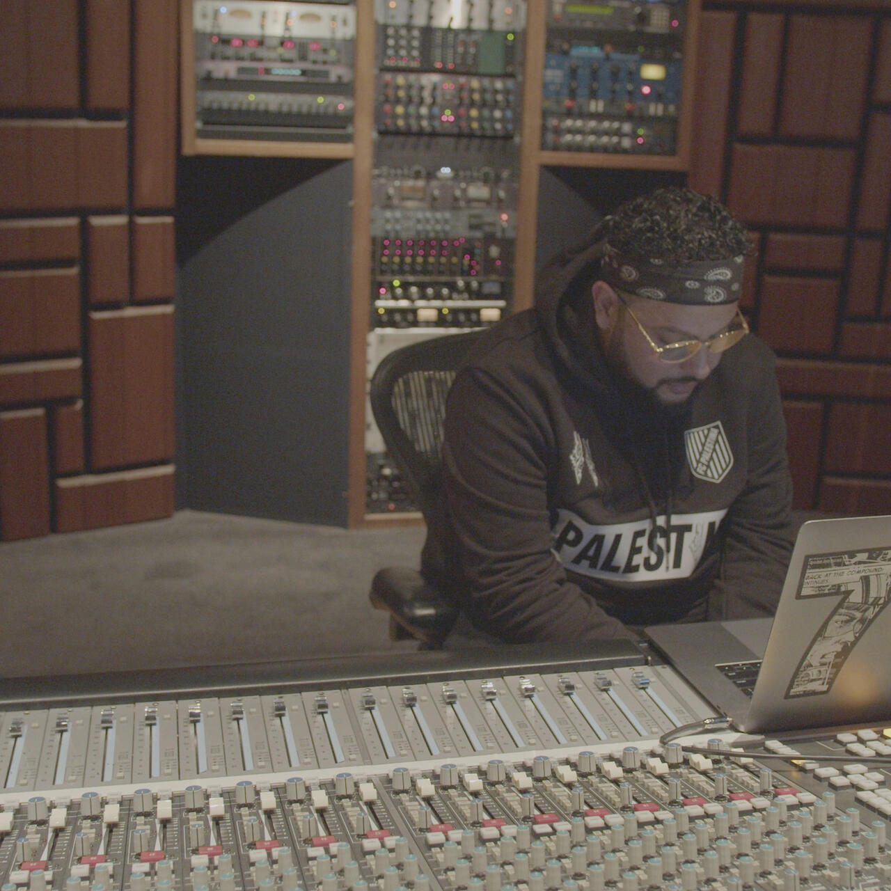 Belly, wearing a headband and a "Palestine" sweatshirt,  sits at his laptop, which is perched on a mixing board in a recording studio.
