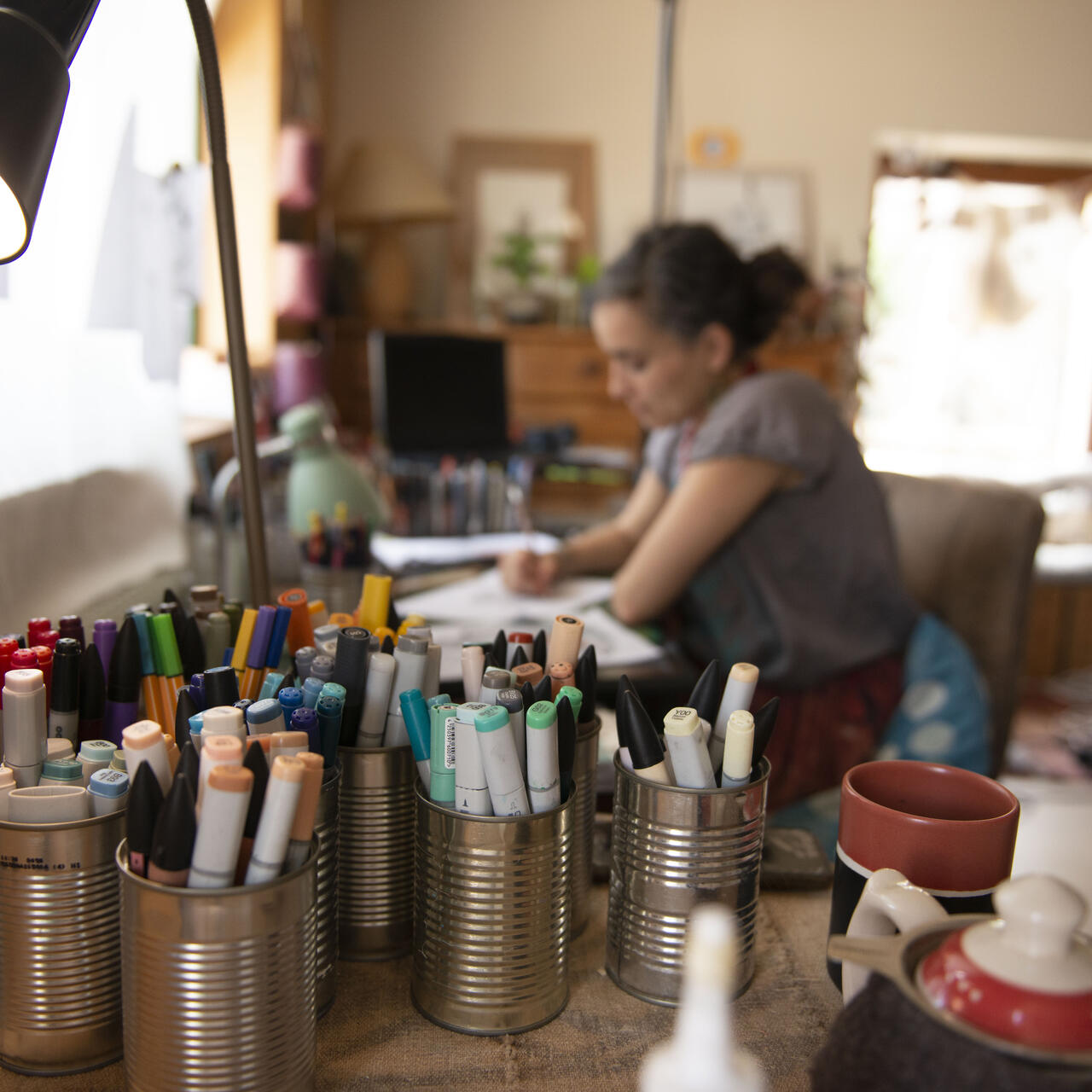 Diala Brisly sits at her drafting table, sketching an illustration for World Refugee Day 2021. Tin cans filled with markers and pens are in the foreground.