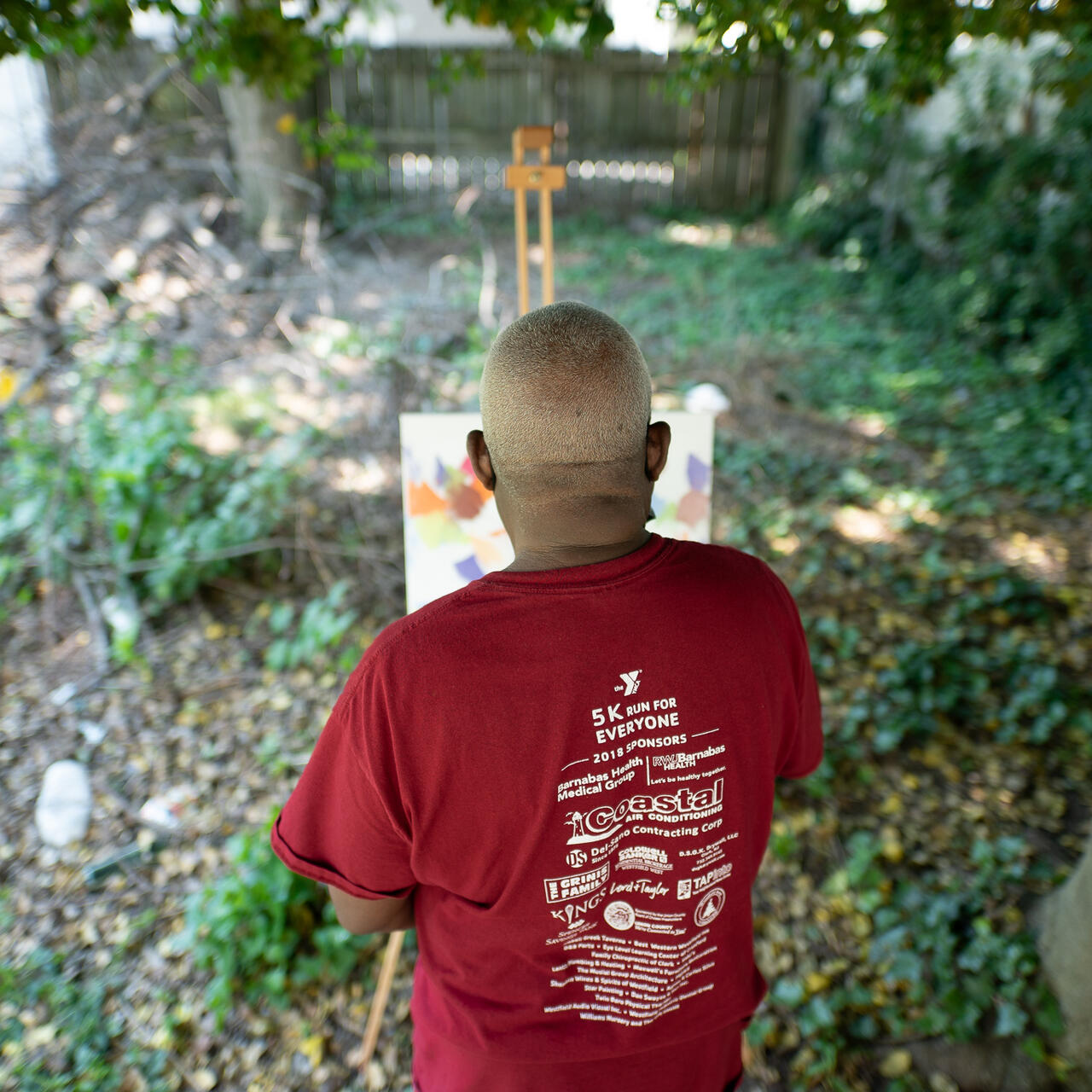 Congolese artist Muyambo Marcel Chishimba, seen from the back wearing a t-shirt, paints an oil painting at an easel in his New Jersey backyard
