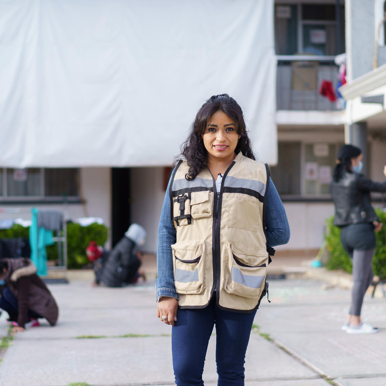 Rosa, wearing a brown vest with large pockets, stands in front of the triage hotel she helps manage in Ciudad Juárez, Mexico. In the background, children draw with chalk on the ground with their parents while other adults converse.