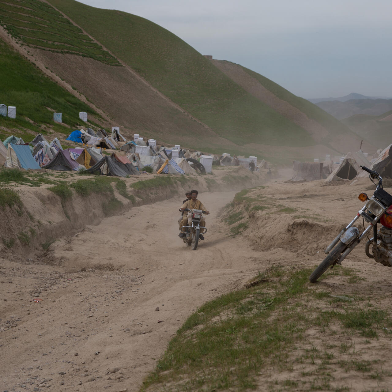 A camp for drought displaced people in Afghanistan. there is a motorcycle in the foreground as well as two men on a motorcycle on the road, and tents in the background on a mountainous landscape.