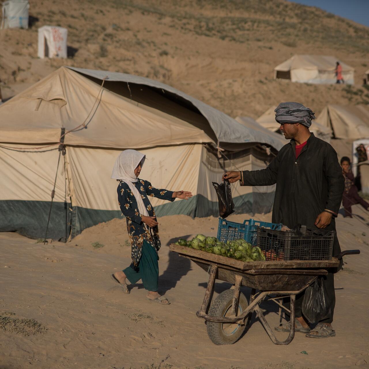 A man pushing a wheelbarrow with vegetables through a tent camp for displaced families in Badghis, Afghanistan hands a girl a plastic bag.