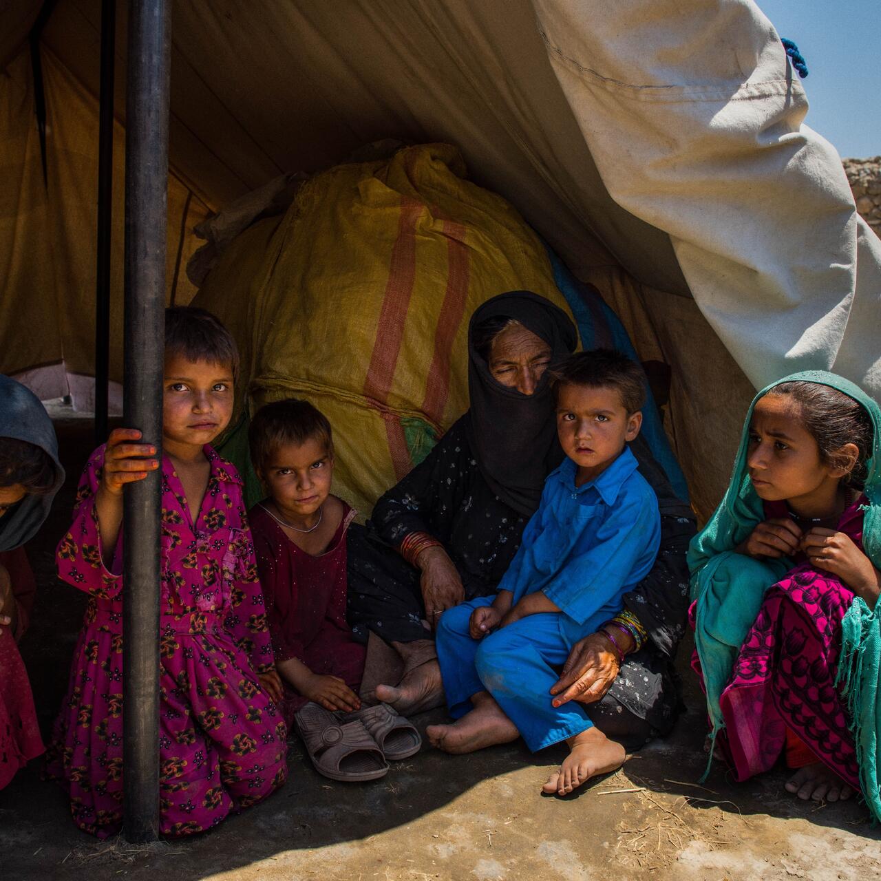 Women and children from an Afghan family living in a displacement camp sit inside the shade of their tent in an arid landscape.