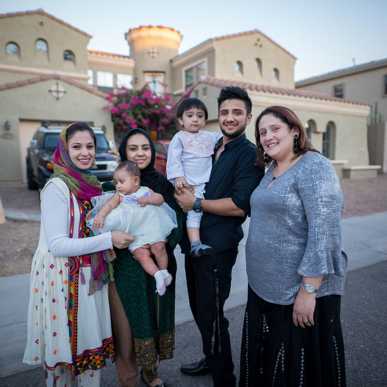 Muska, a woman in her twenties, stands in front of her home with three adult family members, a toddler and a baby.
