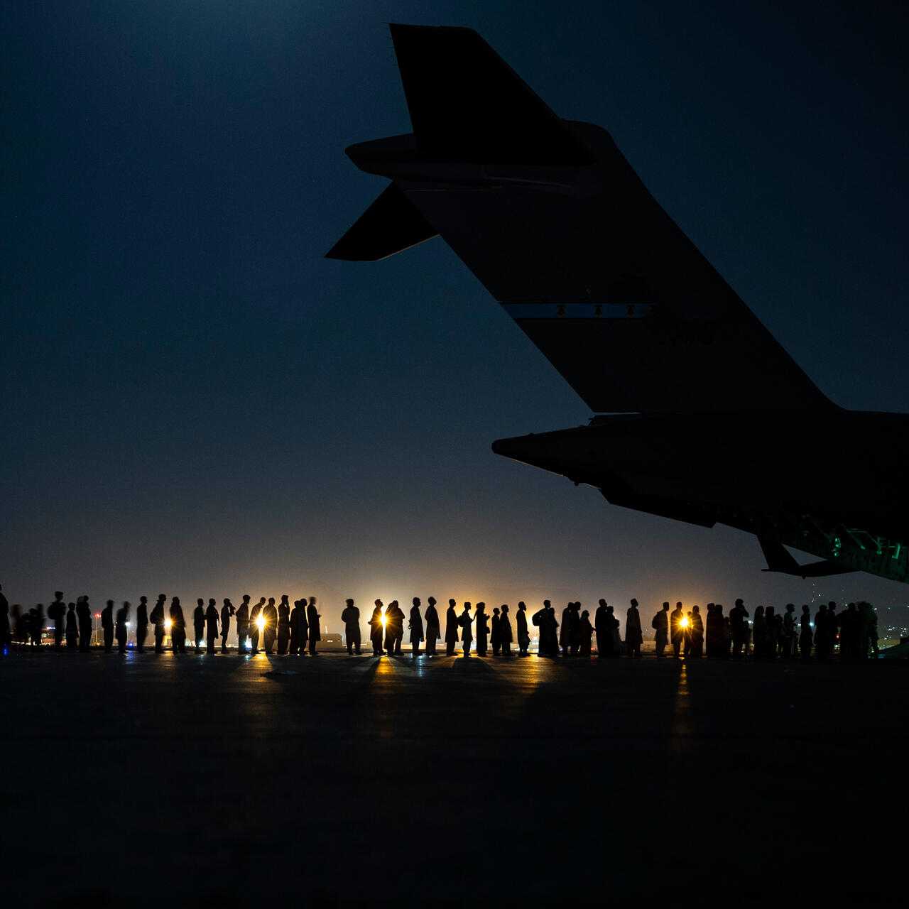 A night shot, with the tail of an airplane in the foreground and a line of people in the background. We can see a light in the distance and just the silhouette of the plane and people. 