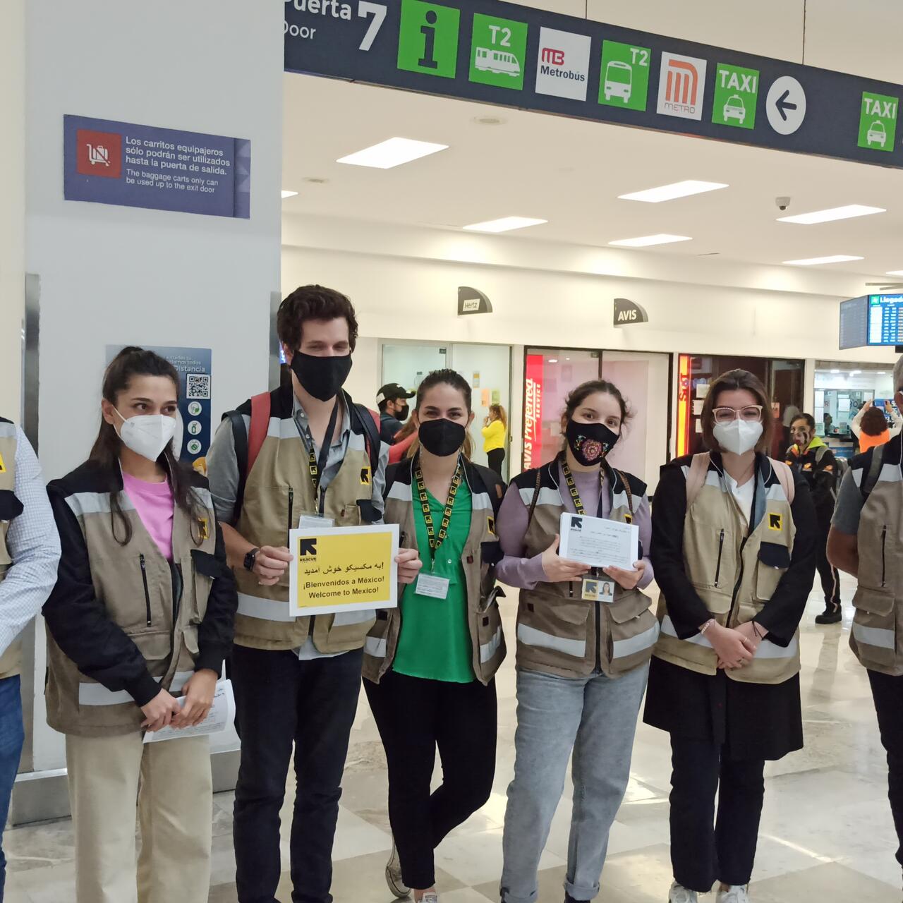 IRC staff members, all wearing masks and IRC-branded vests, stand in a line in an airport terminal. Some are holding welcome signs.
