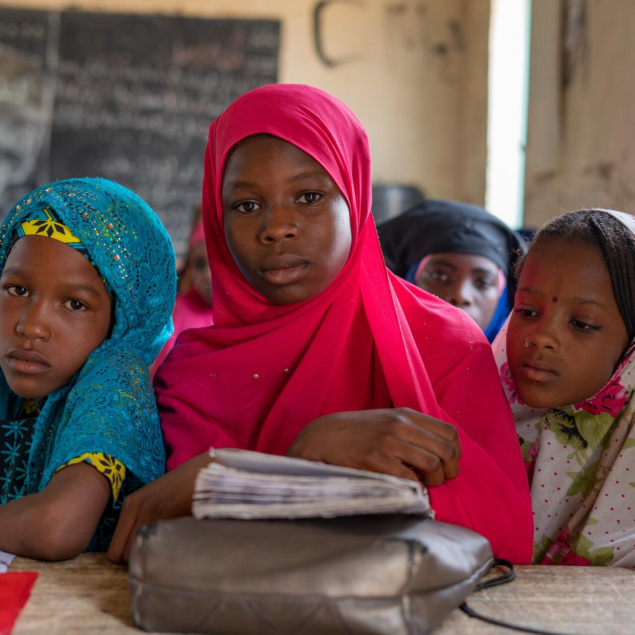 Fifteen-year-old Maryama sits with a serious expression at a school desk with two friends. A school book and papers are on the desk in front of the girls.