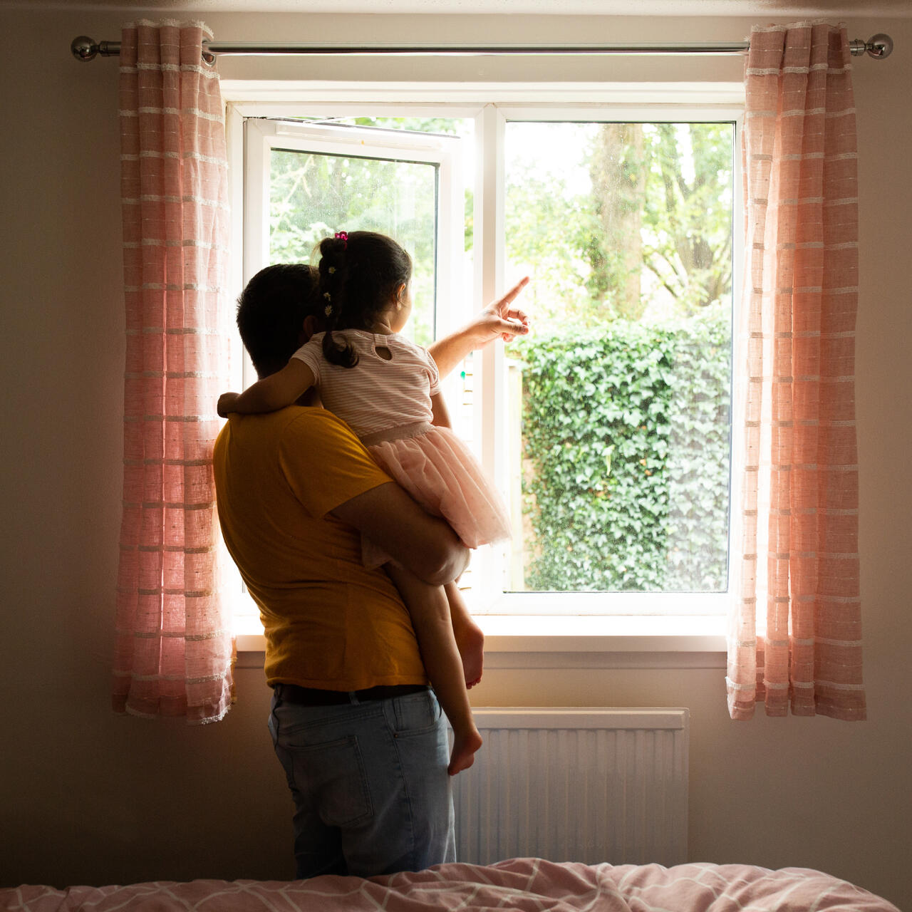 Maasom holds his 4-year-old daughter Nasrin, pointing at a tree as they look through a window at a garden.