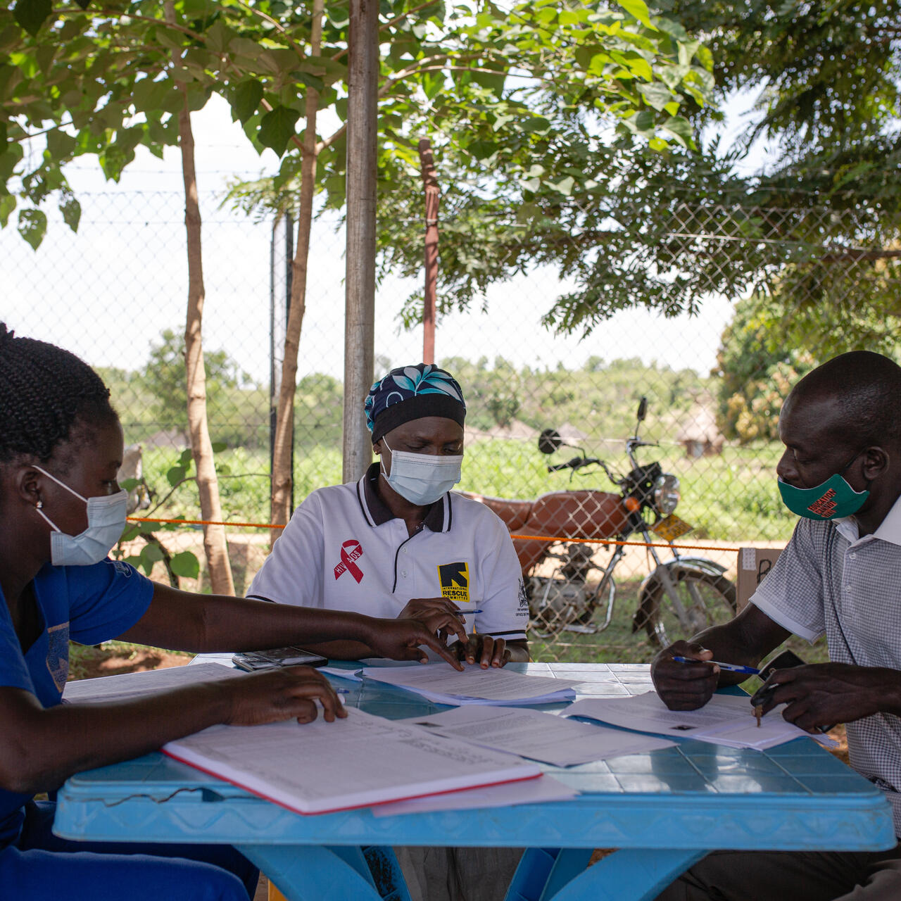Ugandan teacher Azilu Rasulu sits at a table outdoors listening to two female IRC staff provide information about the COVID-19 vaccine.