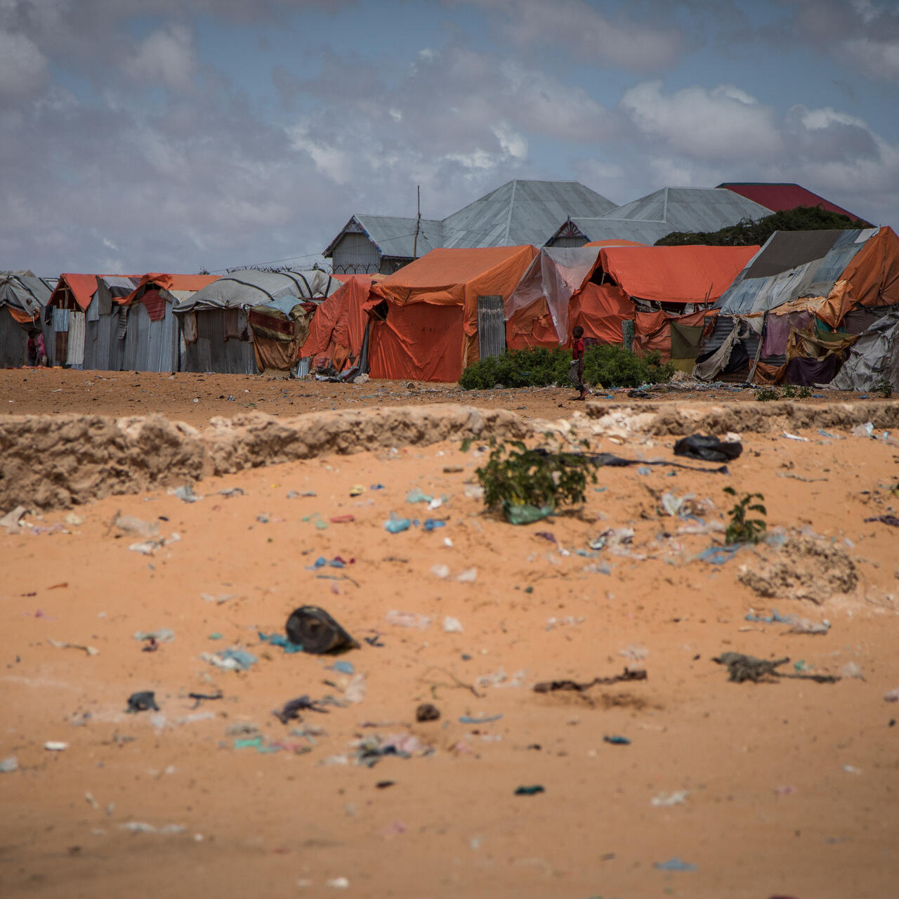 A small boy stands against the backdrop of tents for displaced families in an arid, littered landscape outside Mogadishu, Somalia.
