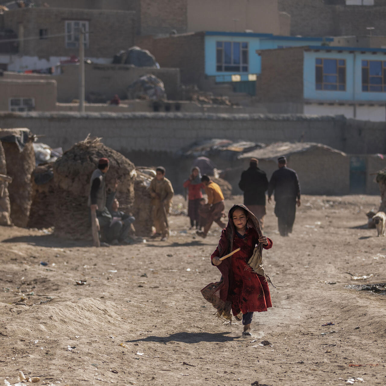 In a camp in Afghanistan, a young girl runs while holding a stick. There are other people and makeshift homes in the background.
