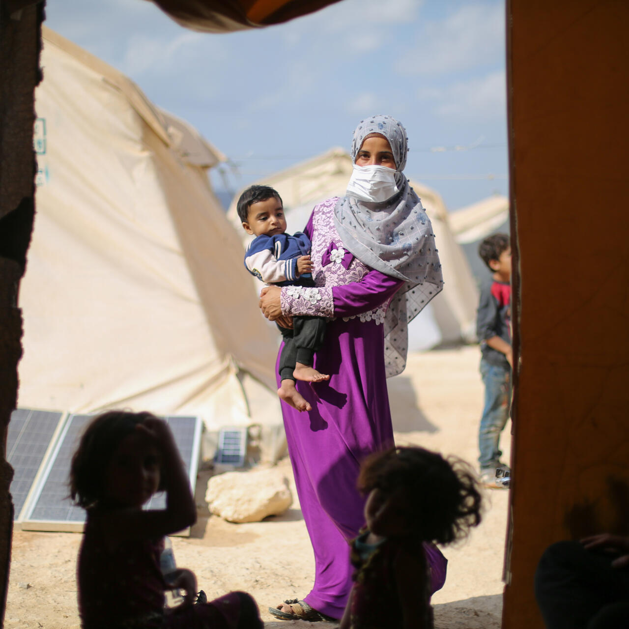 Two children sit on the ground in a tent. Framed in the entrance to the tent is a woman holding a toddler. There are other tents in the background.