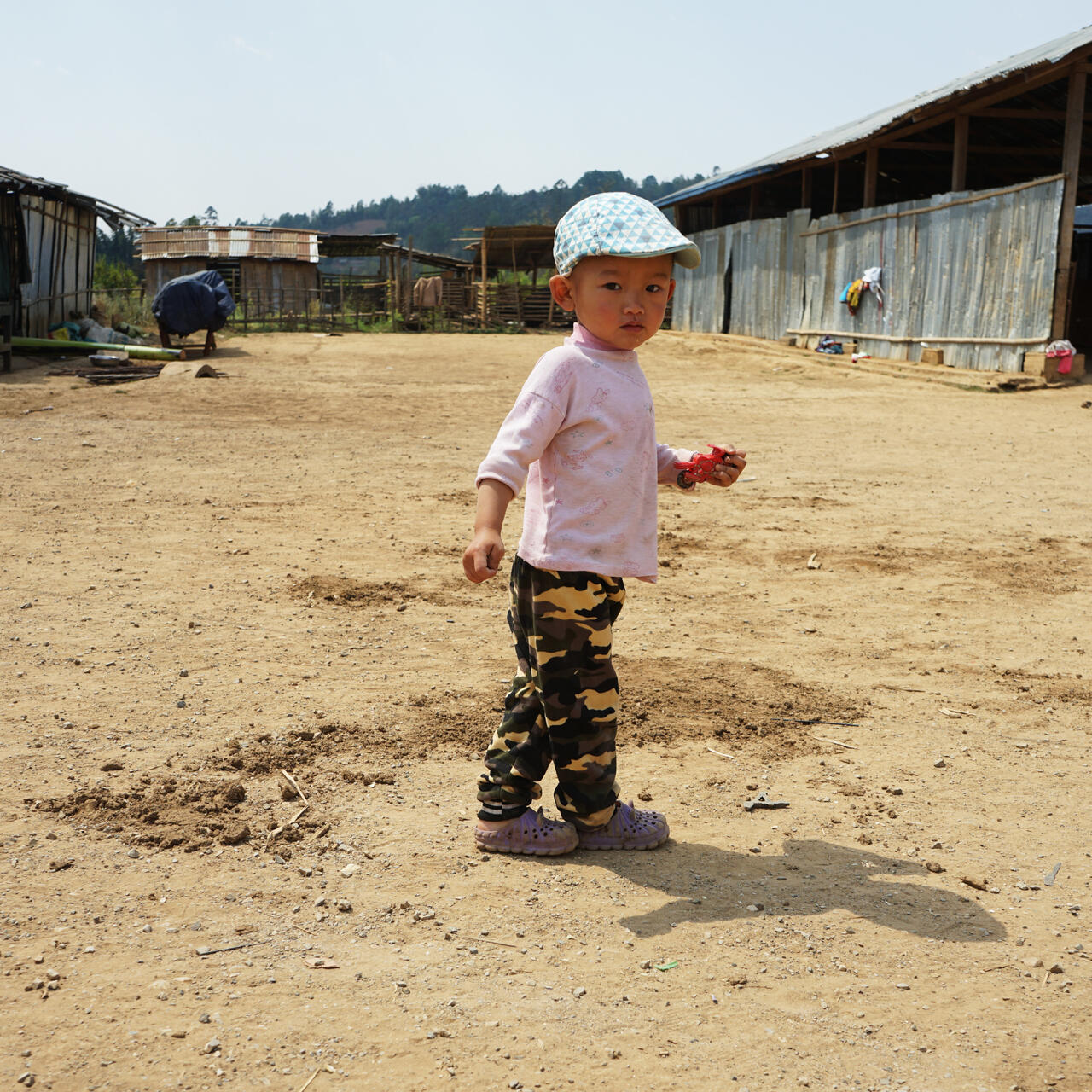 A toddler boy holds a small toy motorcycle as he stands in a wide dusty area between long, corrugated tin structures in a displacement camp.