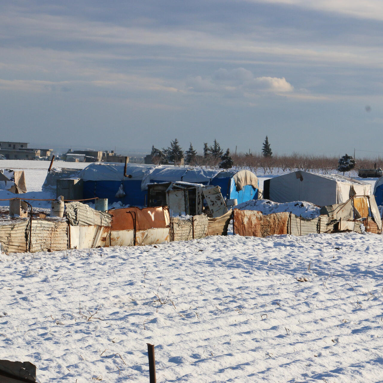 Makeshift shelters housing displaced Syrian families are huddled together in a snow-covered field.