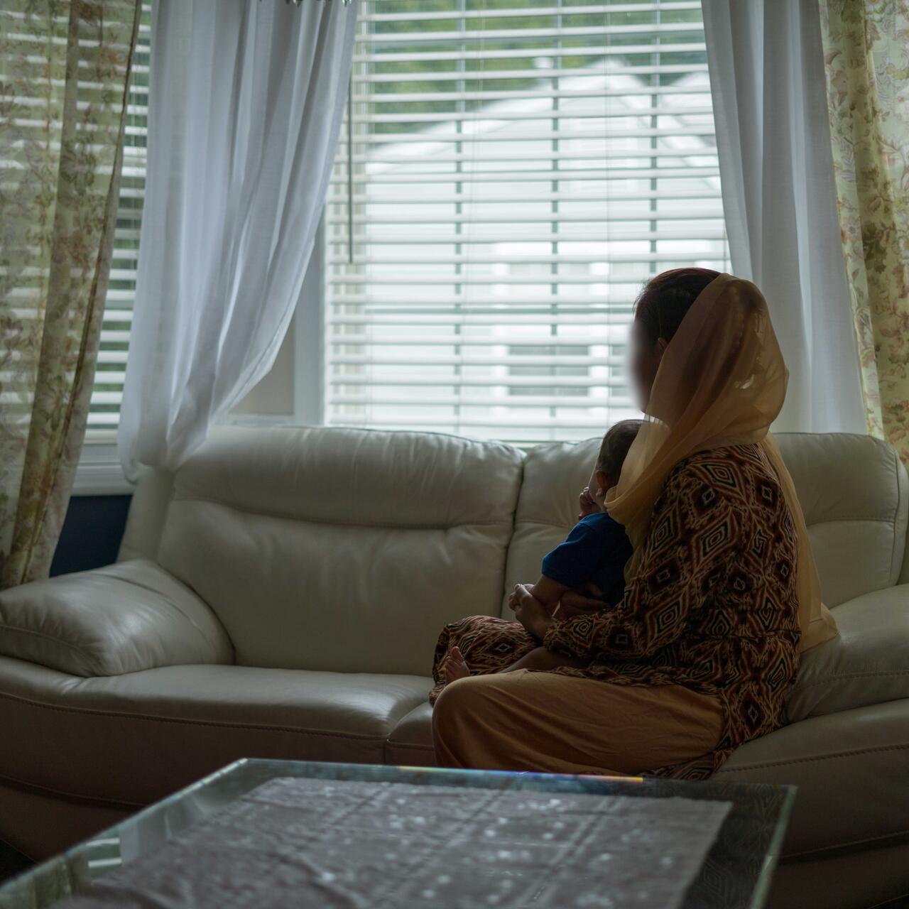 A woman sits on a couch in a living room looking out a window with a toddler on her lap.