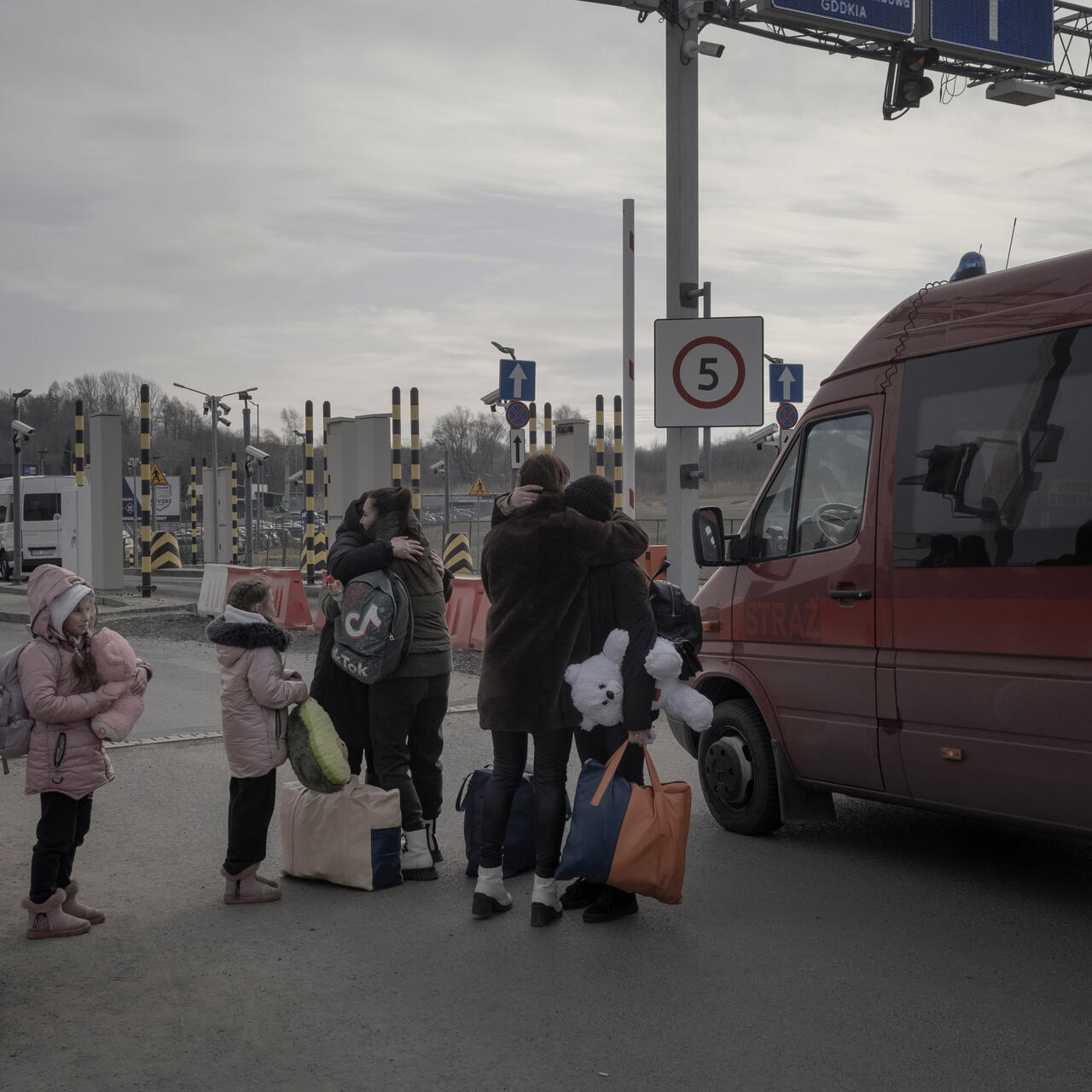 A Ukrainian family embraces at a Ukraine-Poland border crossing next to a van. One person holds a big teddy bear.