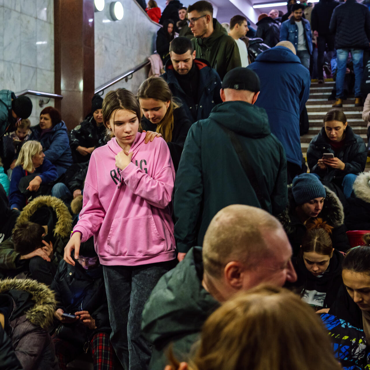 A girl wearing a hoodies stands among people seated on steps as they take shelter in a subway station amid ongoing Russian attacks on cities in Ukraine