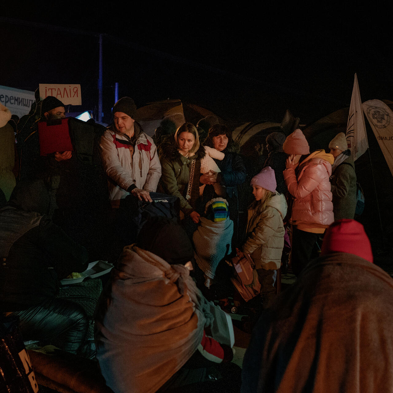 A small group of families with children who fled the war Ukraine, wearing winter coats, stand with their bags on a cold night at the Polish border at Medyka.