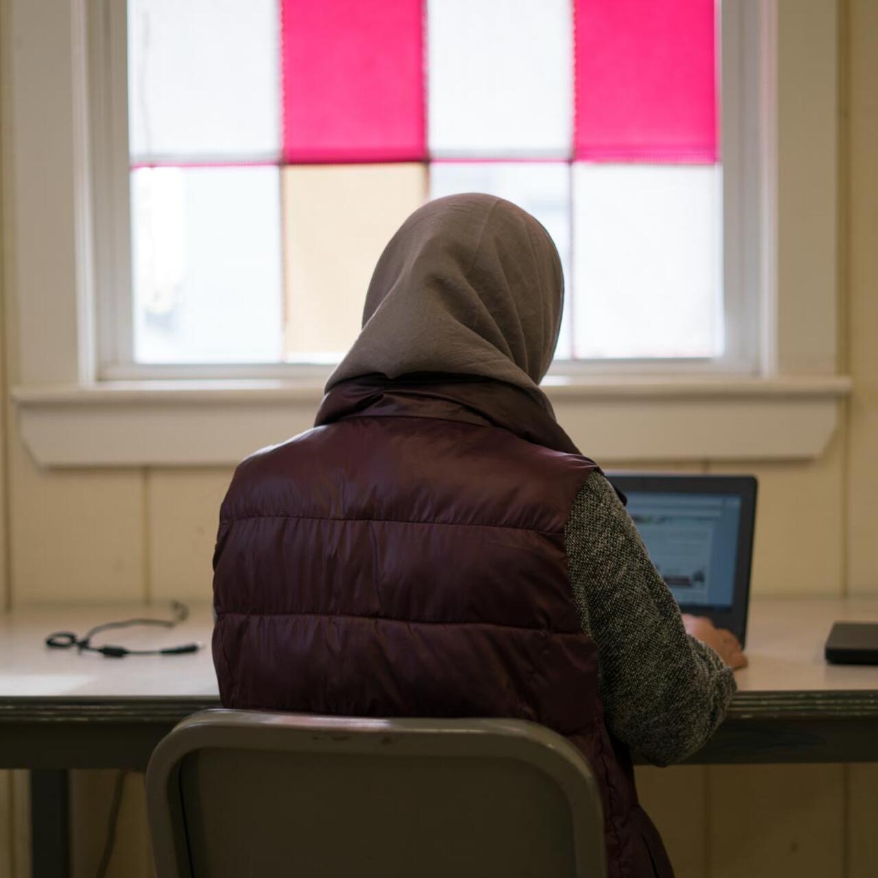 A young woman, with her back to the camera, sits at a desk and types on her laptop.