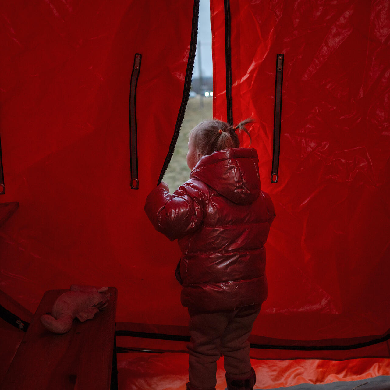 A young girl wearing a red winter coat stands in a red tent, peering out.
