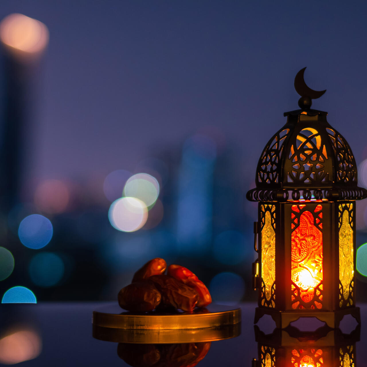 A lantern that has moon symbol on top and small plate of dates fruit with night sky and city bokeh light background for the Muslim feast of the holy month of Ramadan.
