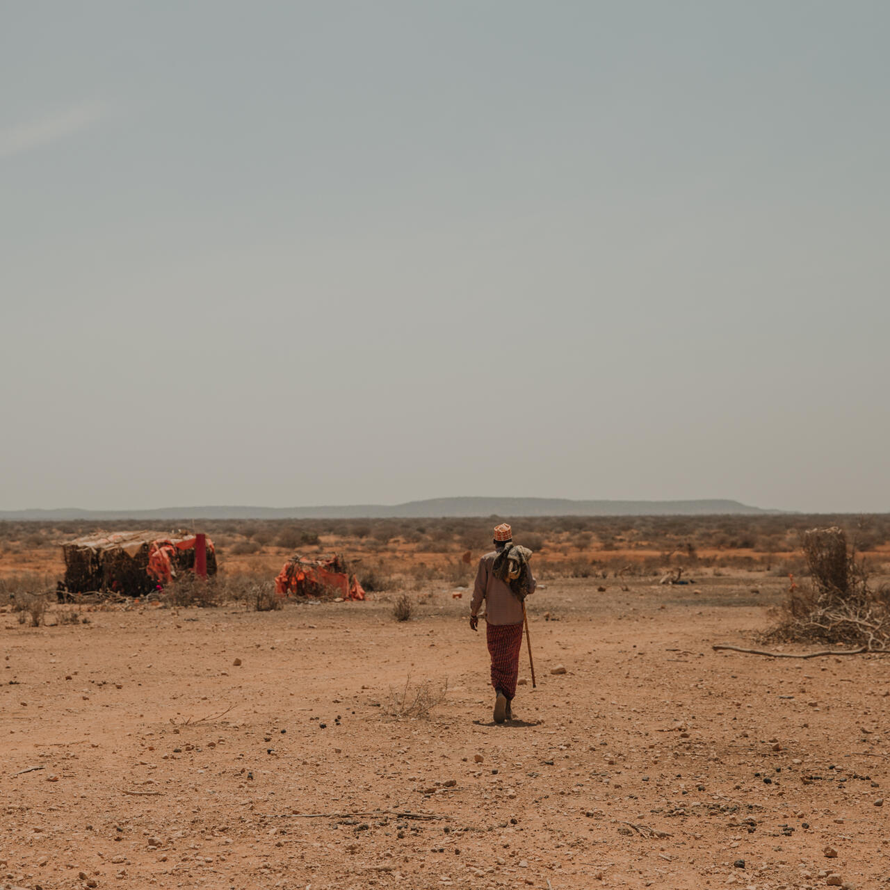 A man carrying a staff walks through a parched landscape in Ethiopia amid makeshift tents with mountains in the distance under a hazy sky.