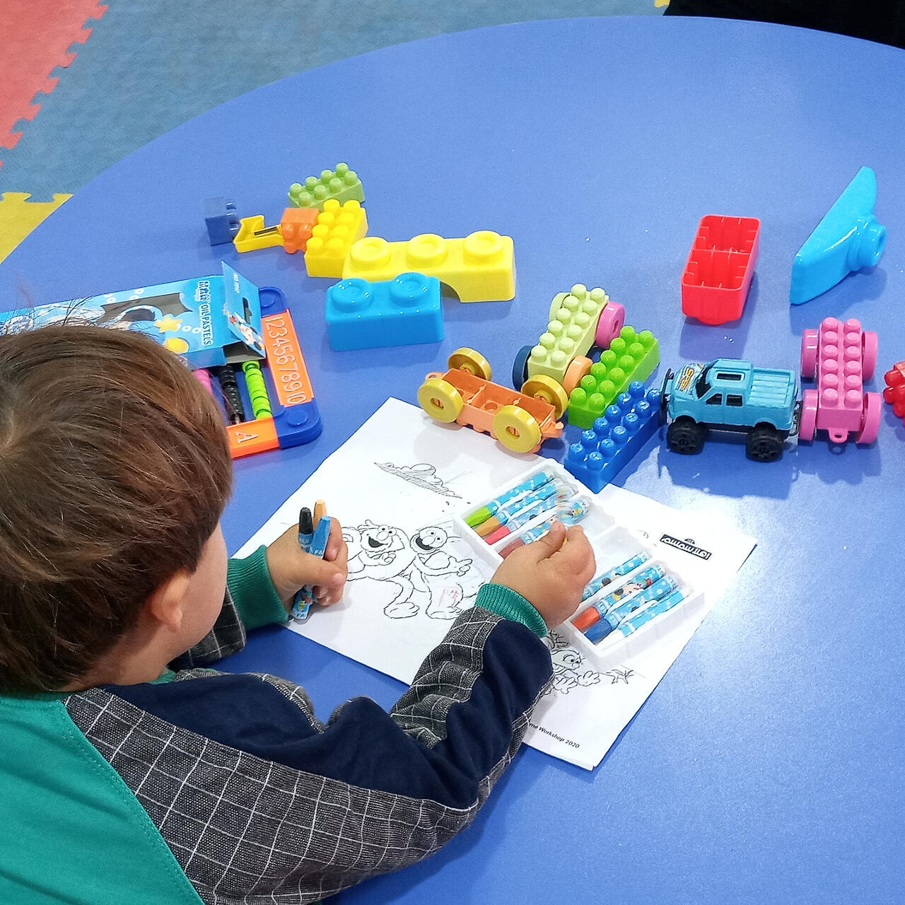 A 5-year-old Syrian boy sits at a low table scattered with toys, choosing oil pastels to color in a page with Sesame Street characters.