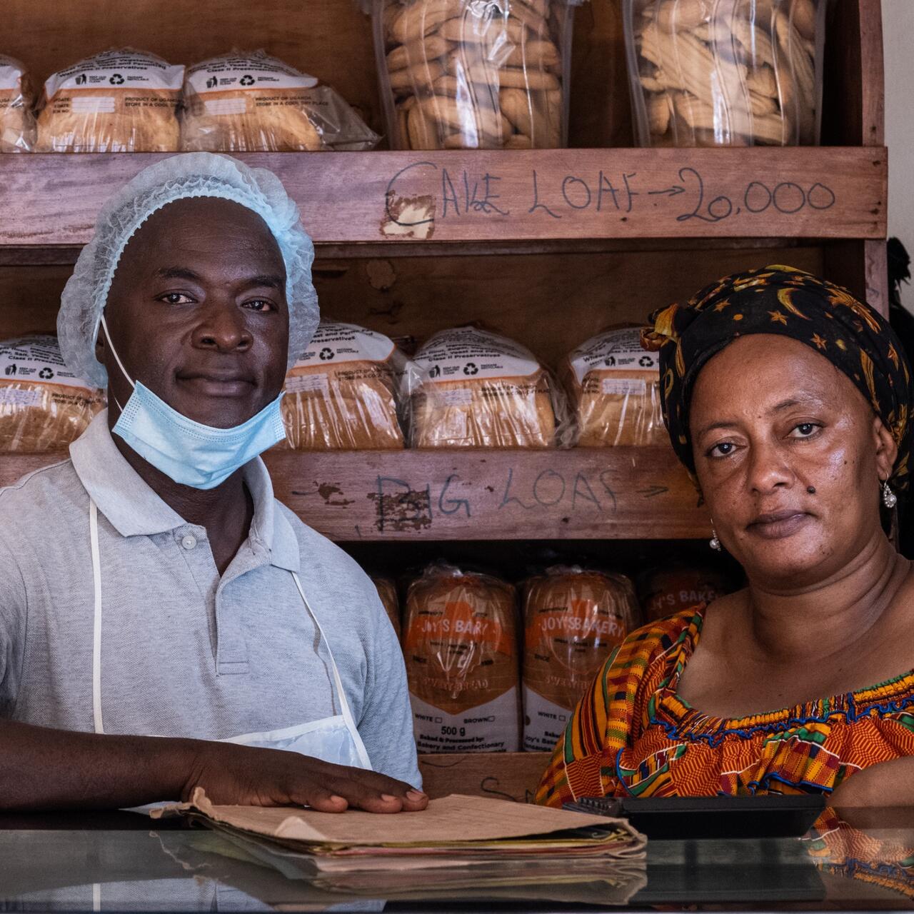 A Ugandan baker wearing a head covering, apron and lowered COVID face mask stands at his bakery counter with his mentee, a woman from South Sudan.