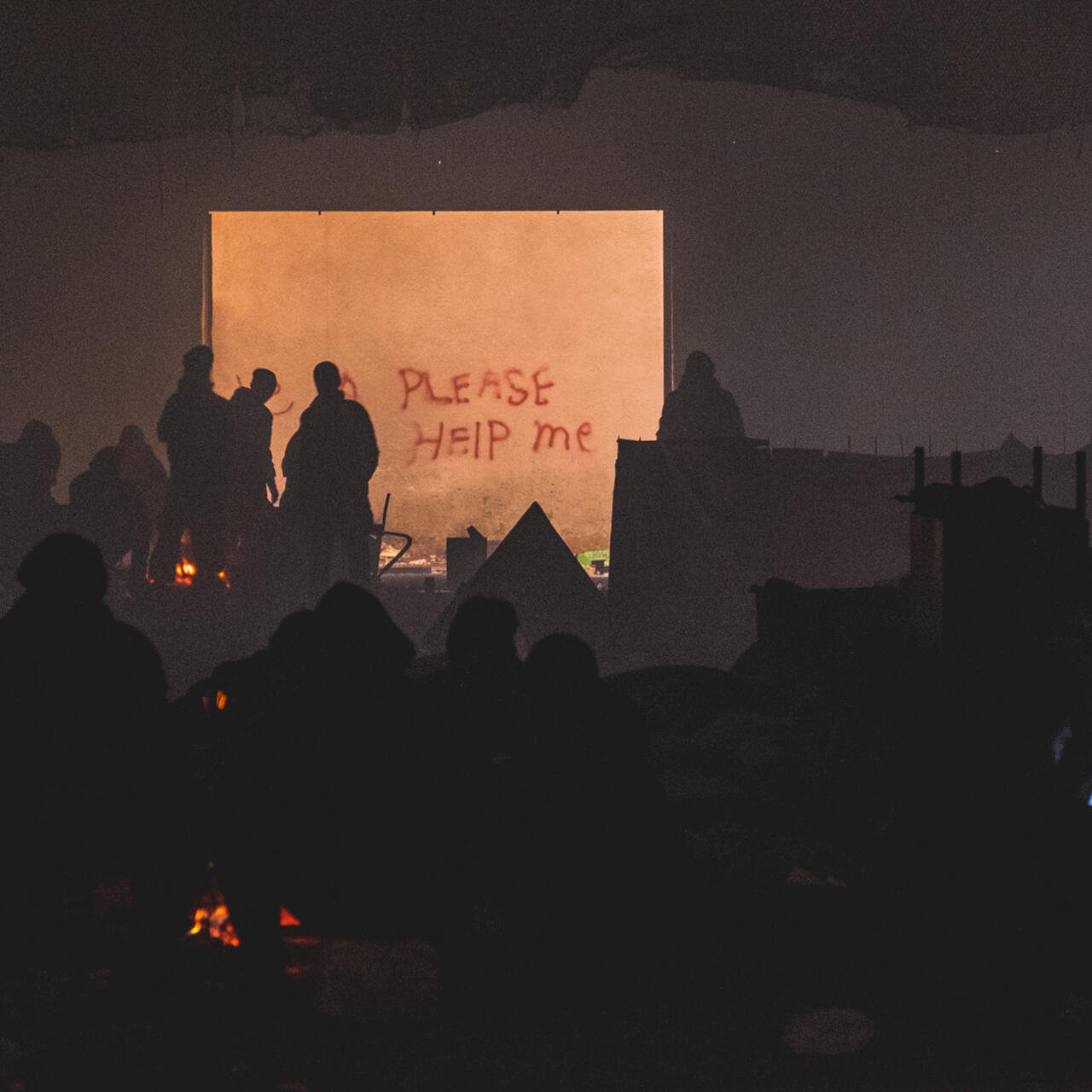 Refugees warm themselves by a bonfire at night on an abandoned industrial site in Belgrade, their figures silhouetted against a slab partly covered by graffiti that reads "PLEASE HELP ME."