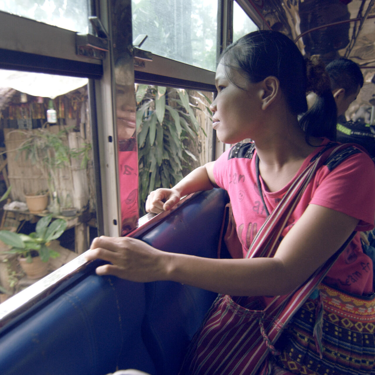 A young woman leaves a refugee camp in Thailand, looking out a bus window.