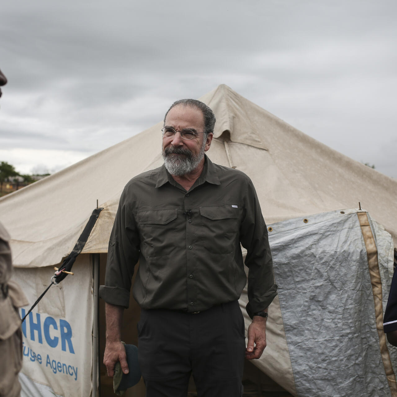 Mandy Patinkin with two IRC aid workers outside a tent in a refugee settlement in Uganda