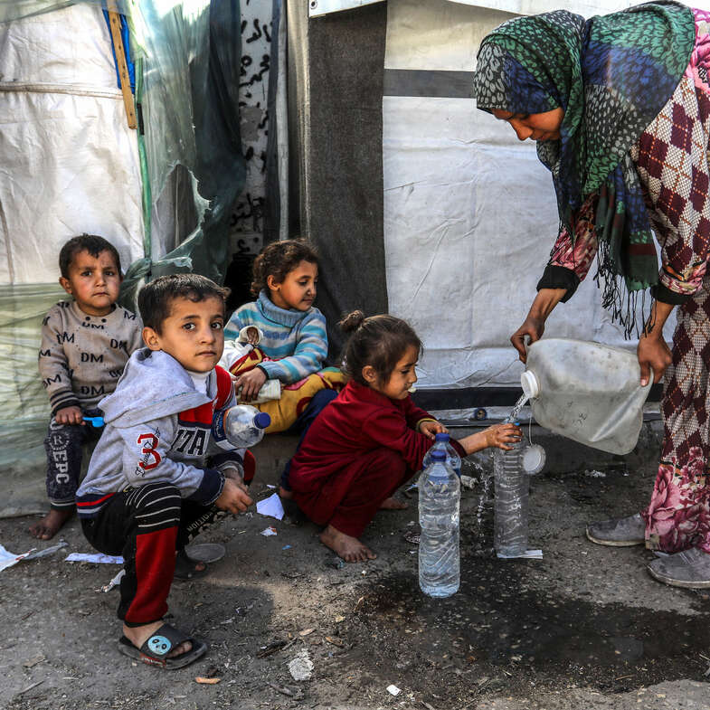 En kvinna i Gaza fyller vatten i flaskor. Barn sitter på gatan.