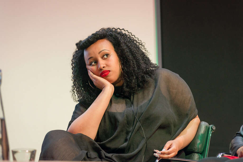 Warsan Shire in a black outfit sitting down, looking right of camera