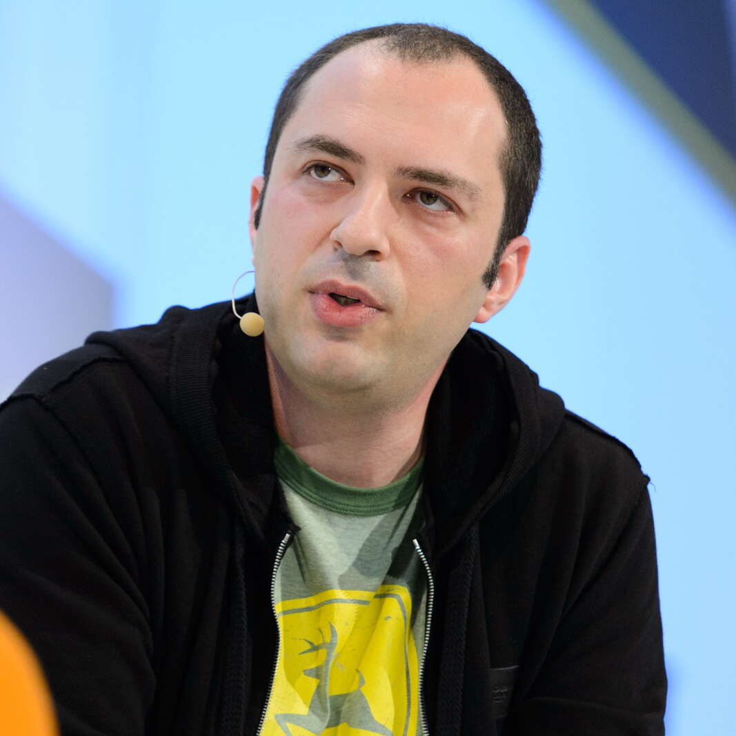 Jan Koum wearing black jacket and green and yellow top, sitting down, speaking with a microphone