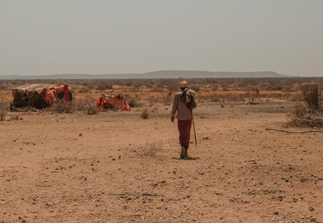 A man carrying a staff walks through a parched landscape in Ethiopia amid makeshift tents with mountains in the distance under a hazy sky.