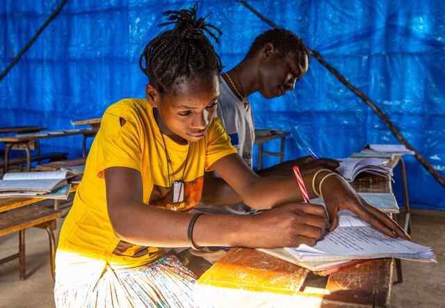 16살의 아사멘치는 교실에 앉아 공부를 하고 있습니다. 그녀는 밝은 노란색 셔츠를 입고 동료 학생 옆에 앉아 있습니다.