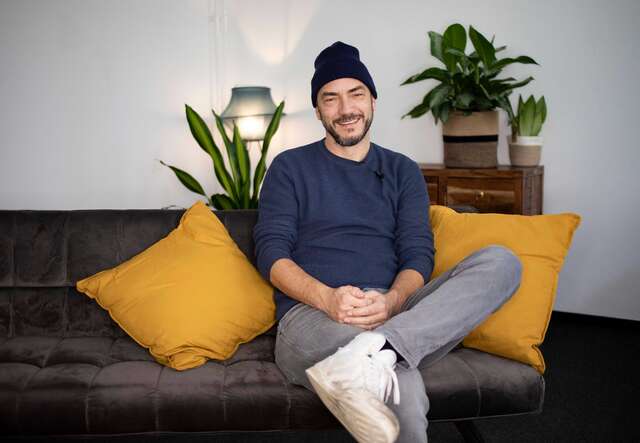 Ein Mann sitzt auf einer Couch und lächelt in die Kamera.