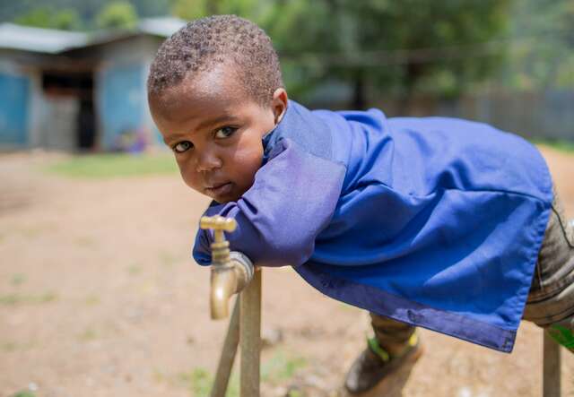 파란색 셔츠를 입은 어린 소년이 수도관 위에 기대어 사진을 찍기 위해 포즈를 취하고 있습니다.
