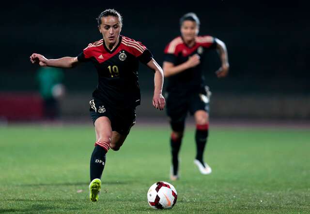 Zwei Fußballspielerinnen in schwarzen Trikots mit roten Streifen rennen auf ein Fußballfeld, wobei eine den Ball kontrolliert.
