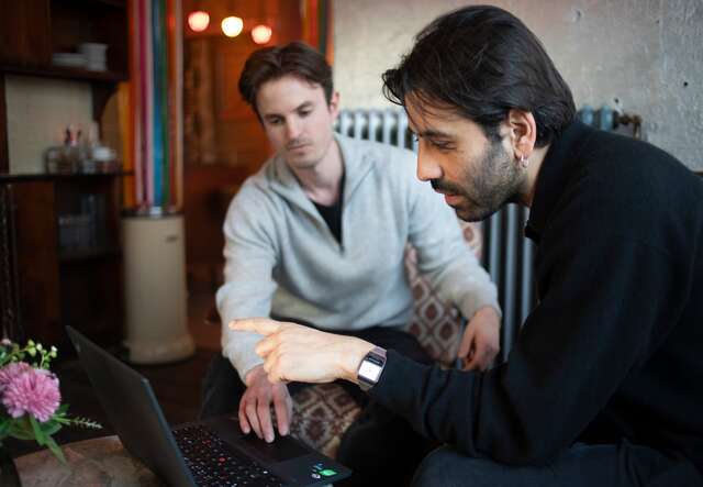 Zwei Männer sitzen auf einem Sofa, lächeln und schauen in die Kamera, während vor ihnen ein Laptop auf einem kleinen Tisch steht.